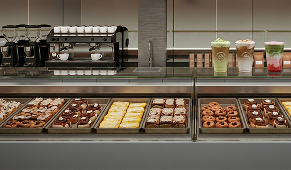 Cinnavibes A bakery display case with assorted pastries, doughnuts, and desserts—including tempting cinnamon rolls—alongside three iced drinks and a coffee machine in the background, capturing the inviting atmosphere of a Cinnamon Desserts Café with true Cinnavibes. Cinnamon Rolls