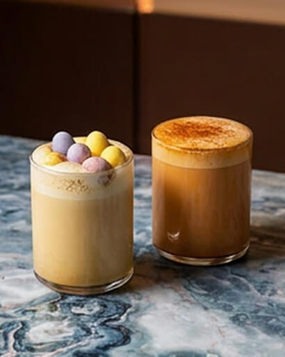 Cinnavibes Two glasses with coffee drinks on a marble table; one topped with colourful round sweets, the other dusted with a layer of cinnamon or cocoa powder—perfect for enjoying at Cinnamon Deserts Café, where the classic aroma of cinnamon rolls and signature Cinnavibes fill the air. Cinnamon Rolls