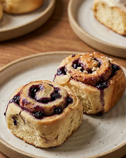 Cinnavibes Two blueberry swirl rolls sit on a beige ceramic plate, echoing the sweet artistry found at Cinnamon Deserts Café. In the background, another plate holds a partially eaten roll—perfect for anyone craving those signature Cinnavibes and hand-crafted cinnamon rolls. Cinnamon Rolls