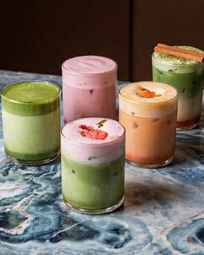 Cinnavibes Five colourful iced drinks in clear glasses are arranged on a blue marbled surface at Cinnamon Deserts Café. Drinks include green, pink, and orange hues, with various toppings like fruit, cinnamon, and foam—each sip capturing the sweet Cinnavibes that pair perfectly with their signature cinnamon rolls. Cinnamon Rolls